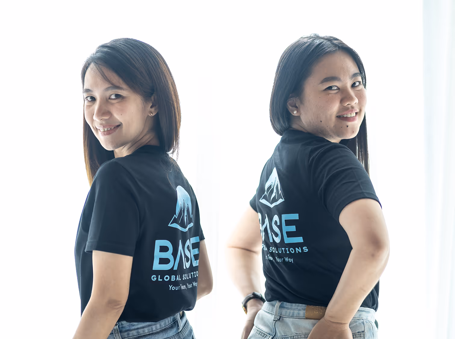 Two smiling women standing back-to-back wearing black BASE Global Solutions t-shirts, with the company logo and slogan 'Your Team, Your Way' visible on the back.
