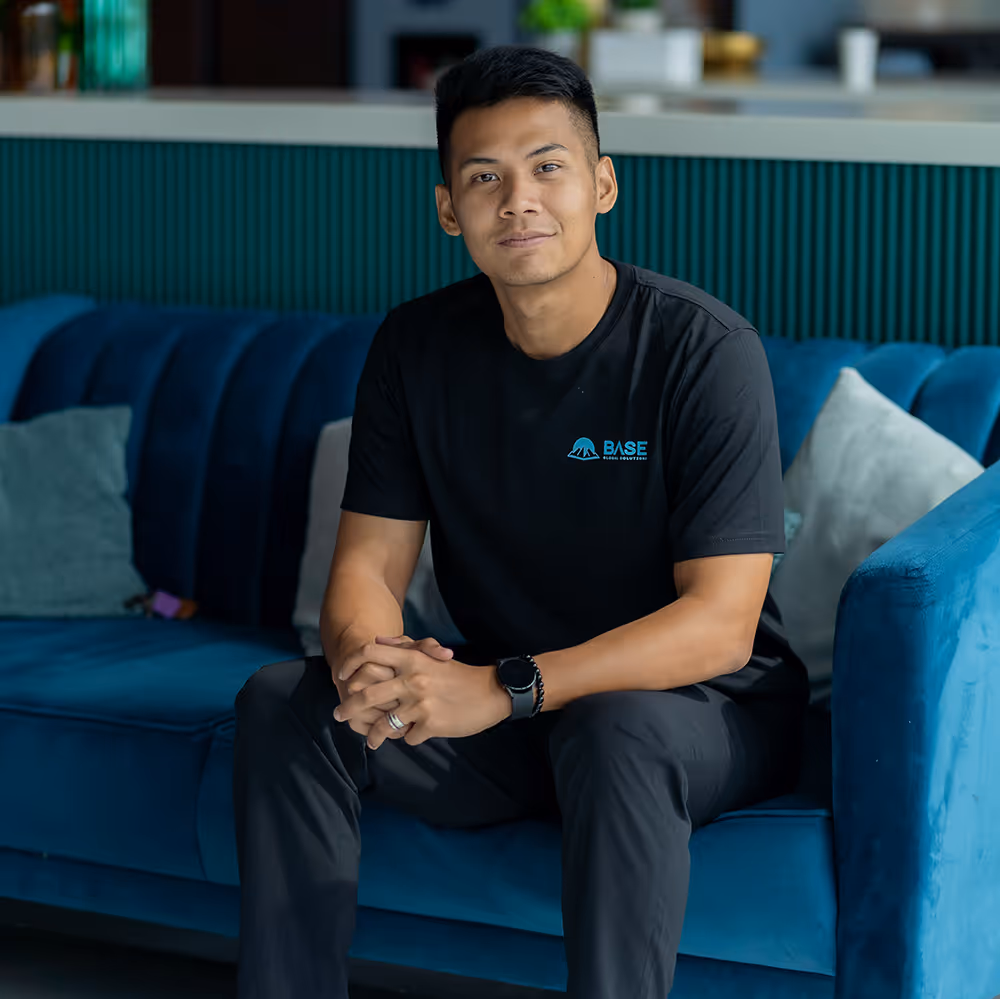 Man wearing a black BASE Global Solutions t-shirt sitting on a blue couch, looking confidently at the camera with hands clasped.