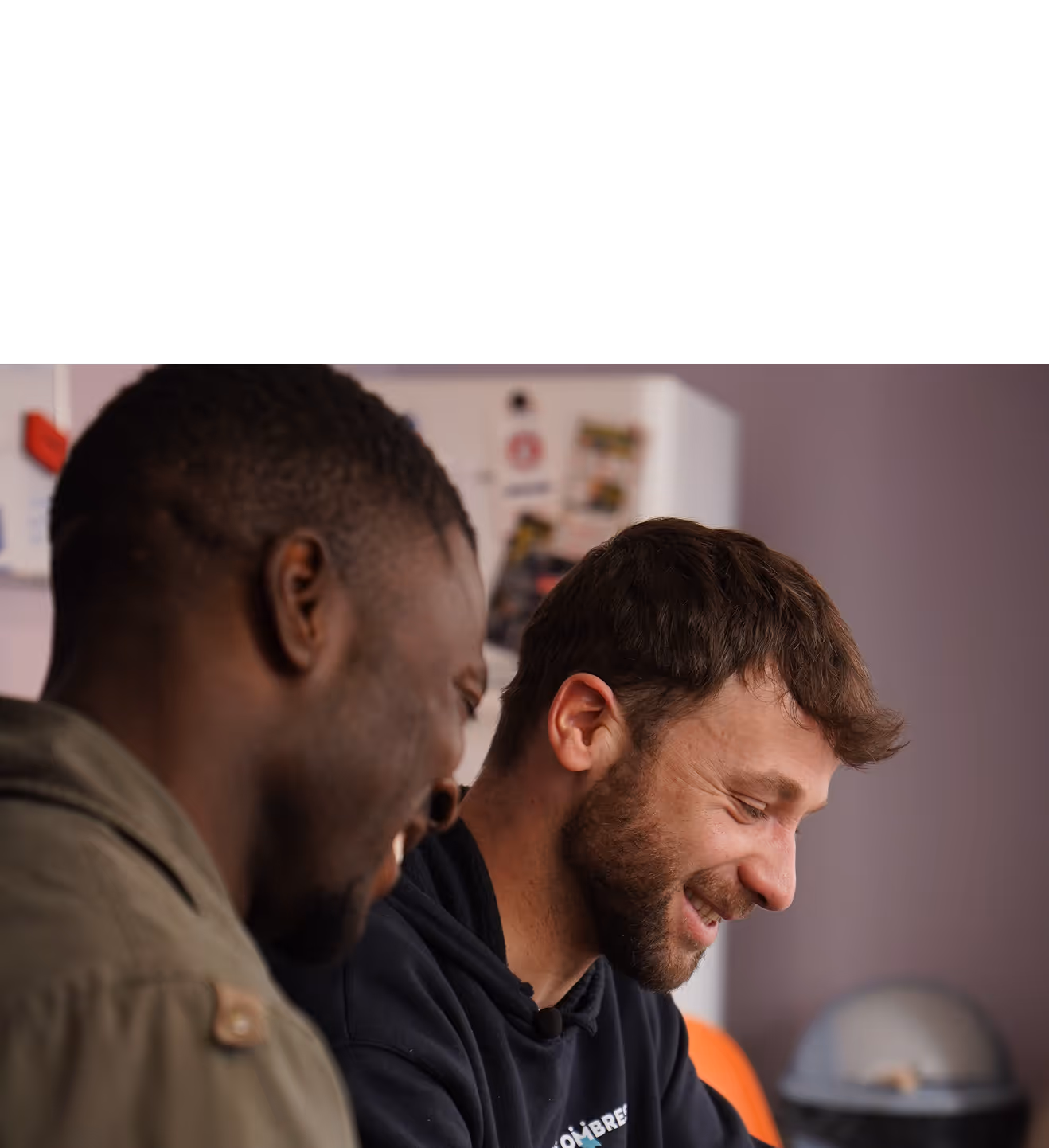 Deux hommes souriants travaillant ensemble à une table avec un ordinateur portable dans une pièce au mur violet.