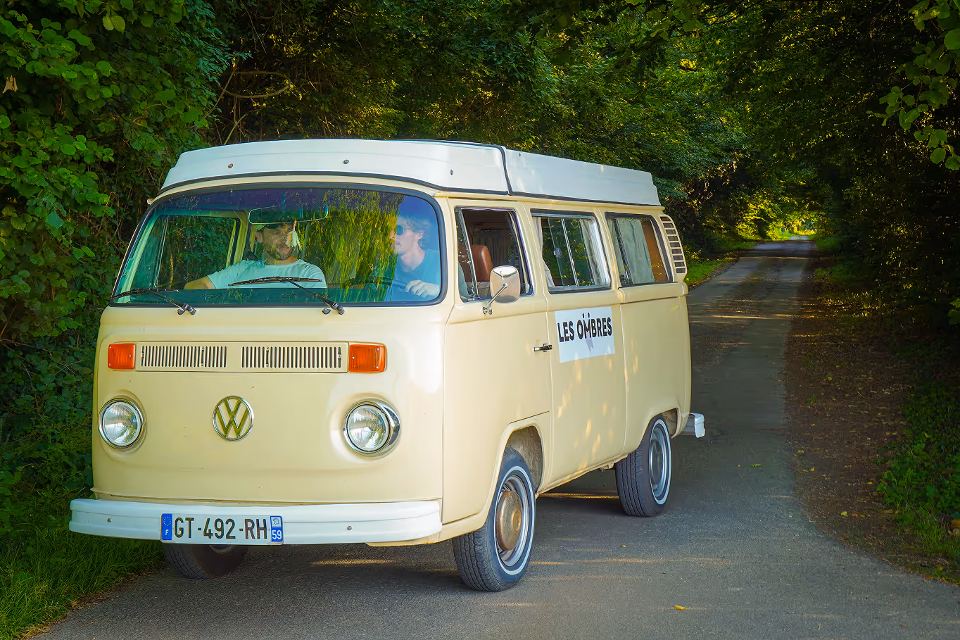 Van Volkswagen beige avec deux personnes à bord sur une route bordée d'arbres, avec un panneau 'LES OMBRES' sur la porte.