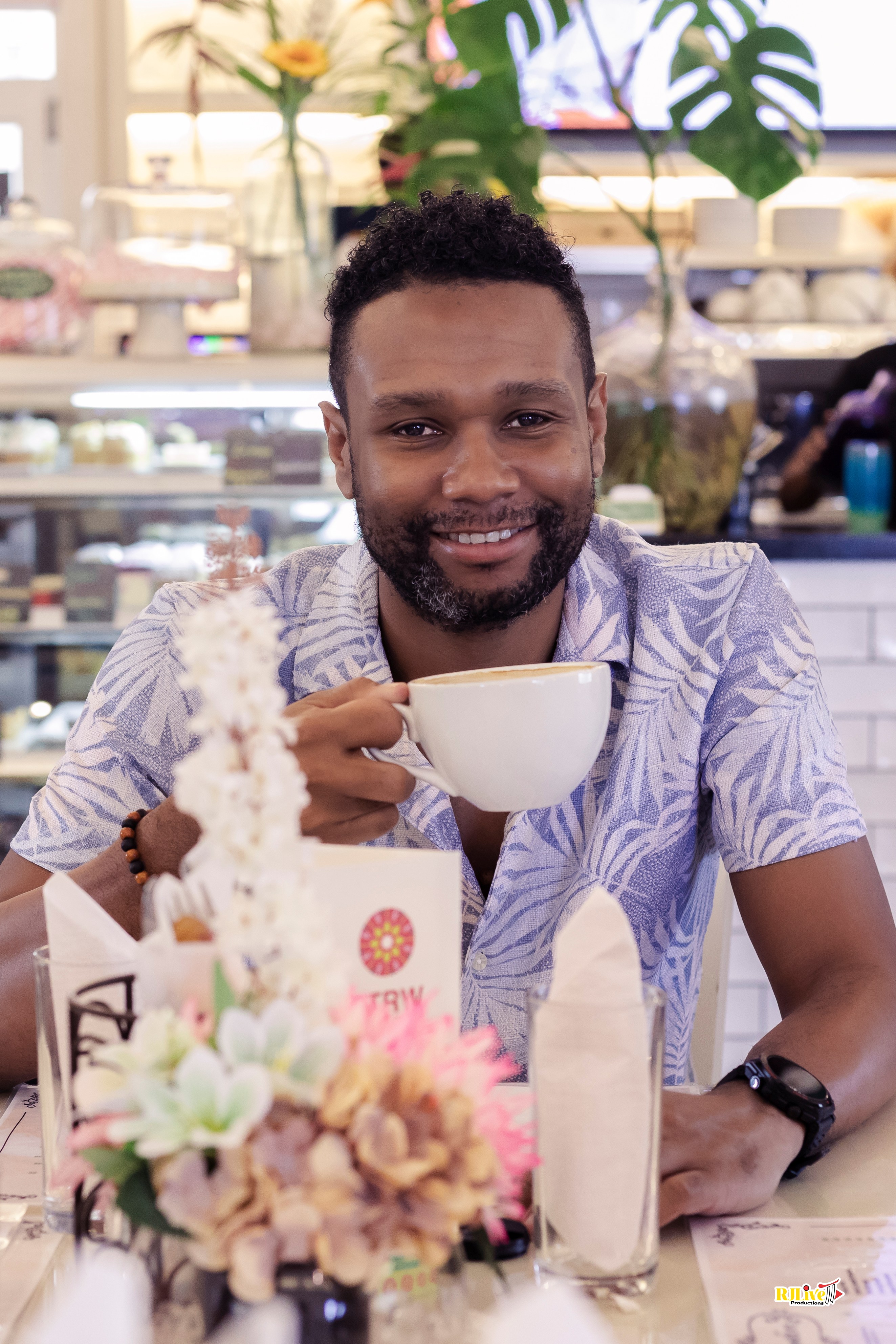 Smiling man with curly hair and beard holding a white coffee cup in a cafe setting with flowers and napkins on the table.