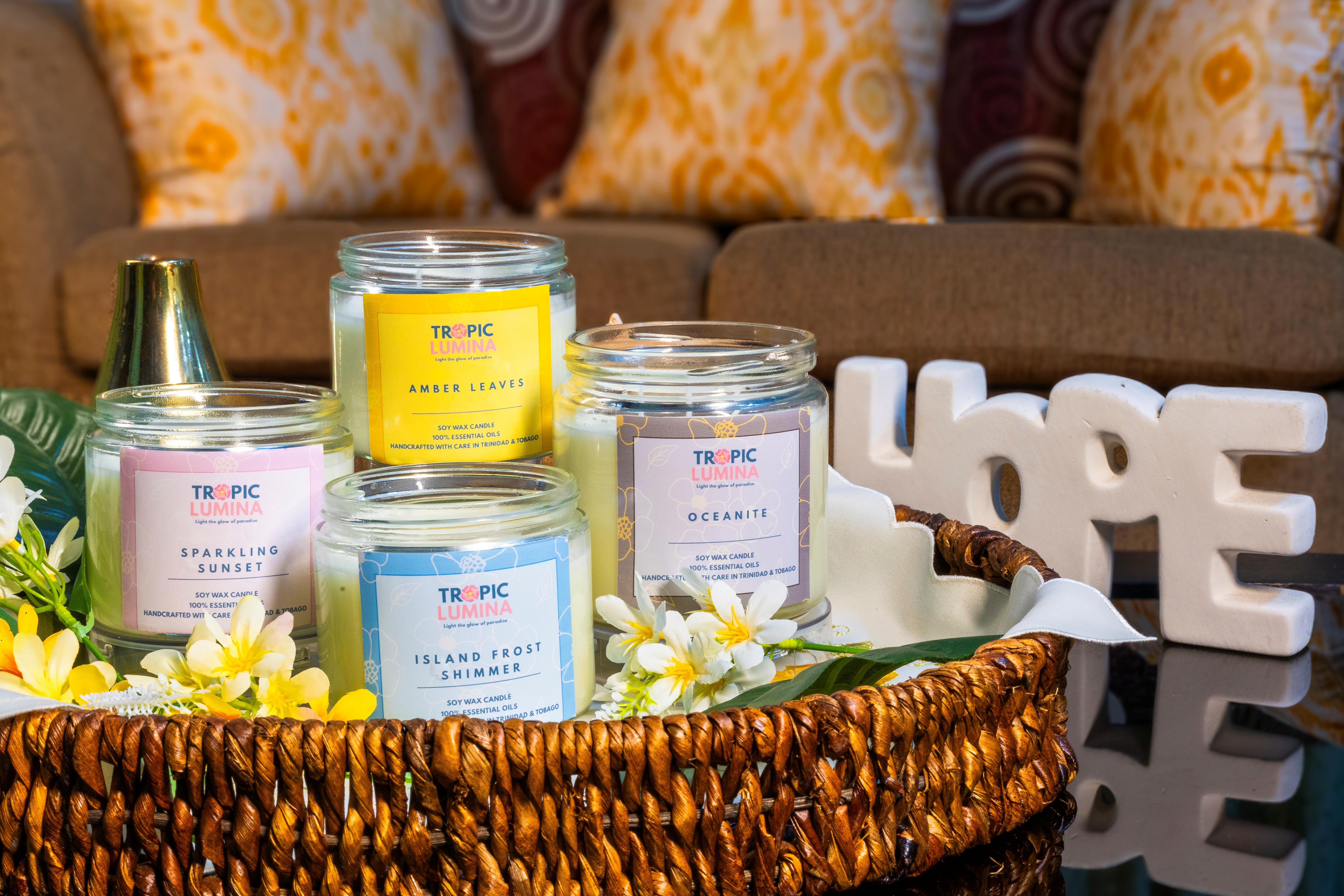 Four Tropic Lumina soy wax candles with labels Amber Leaves, Sparkling Sunset, Island Frost Shimmer, and Oceanite in a woven basket decorated with yellow and white flowers, with a white HOPE sign in the background.