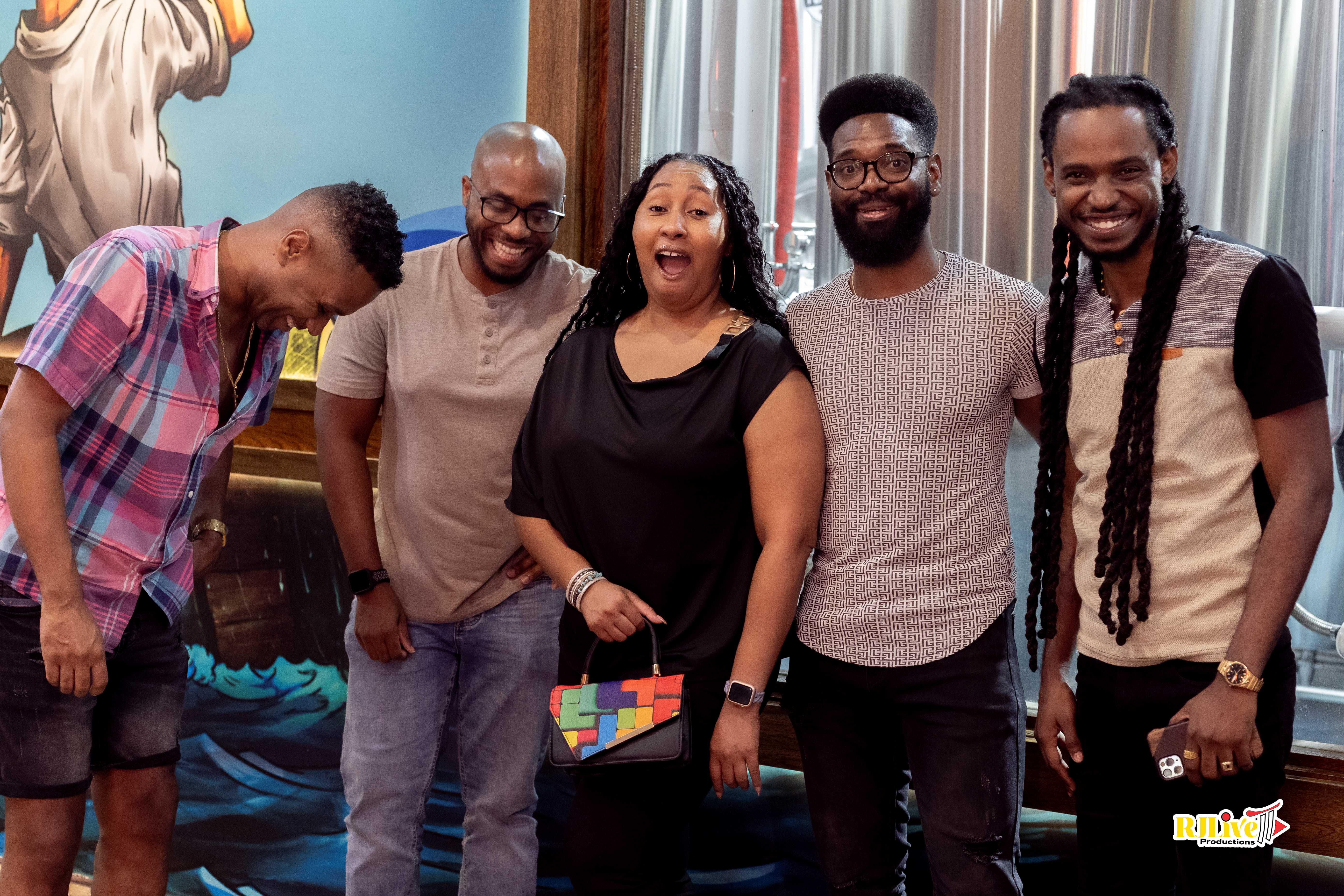 Five people standing indoors smiling and laughing, with a colorful mural and metal tanks in the background.