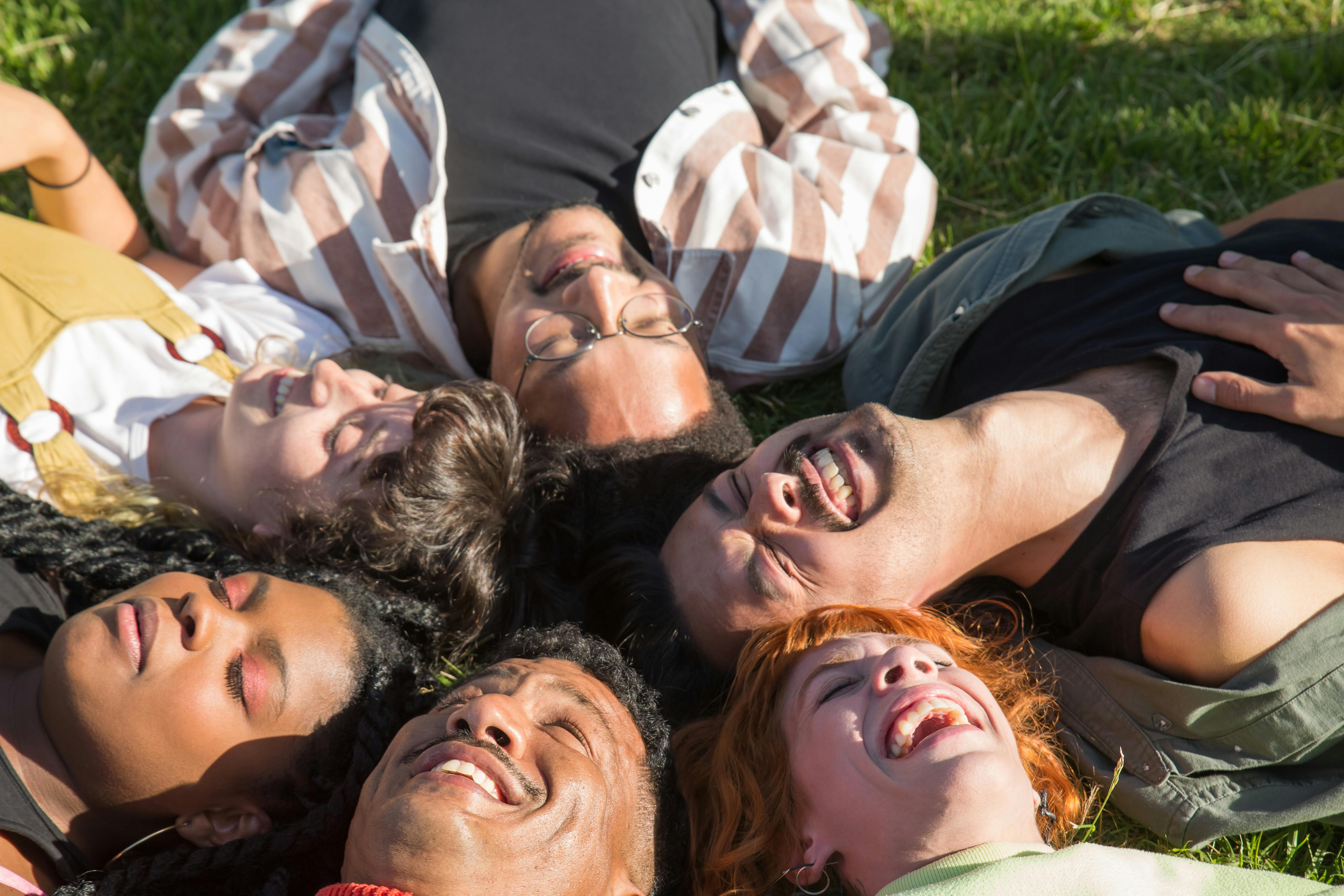 People from different backgrounds lying together on the grass, laughing in the sunlight, symbolizing connection, community, and belonging across cultures.