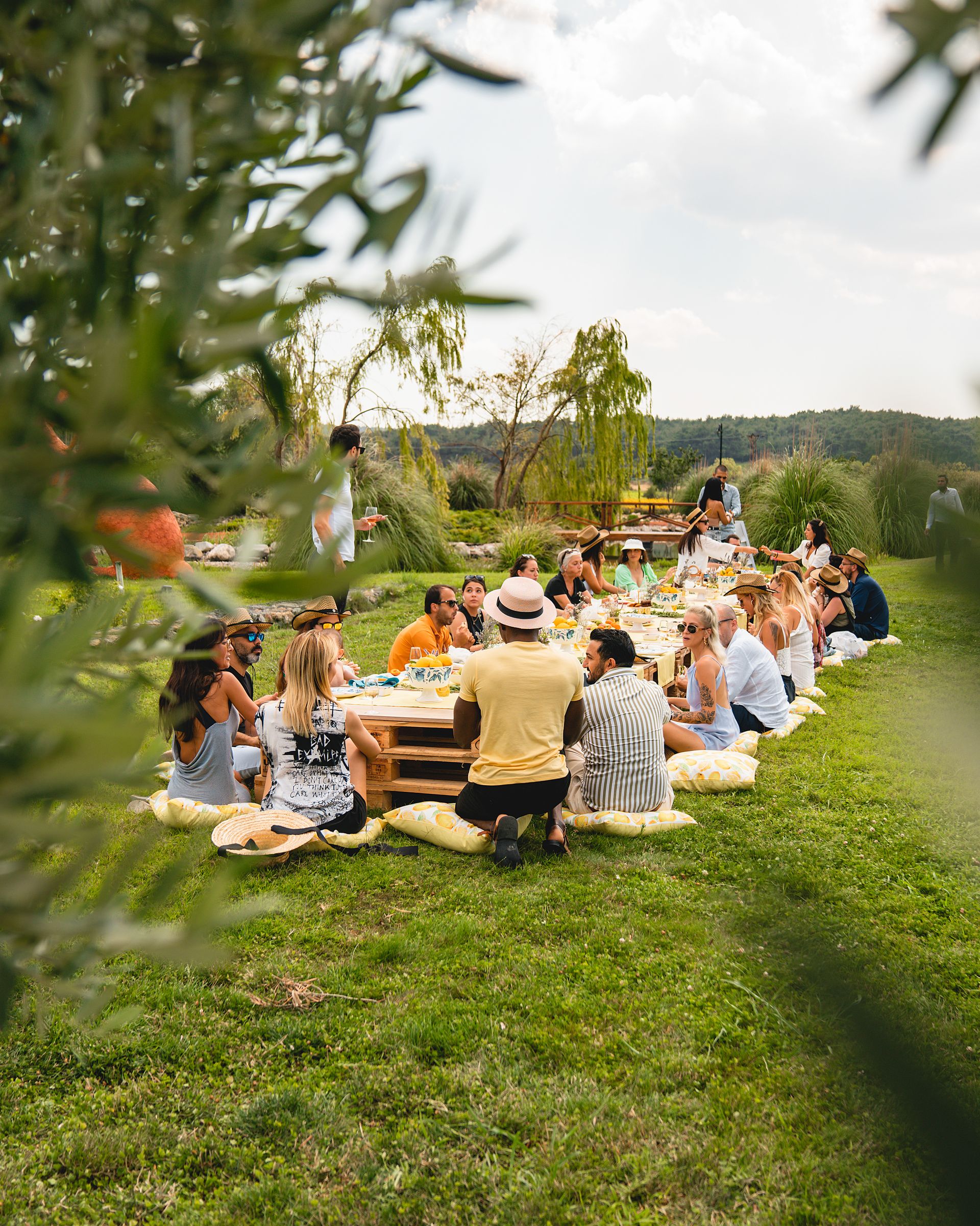 A group of people gather around a long, low dining table set on a grassy lawn for an outdoor meal. Guests sit together enjoying food, conversation, each others' company in a lush garden surrounded by trees, ornamental grasses. Social interaction and activities make people in the group belonging. 