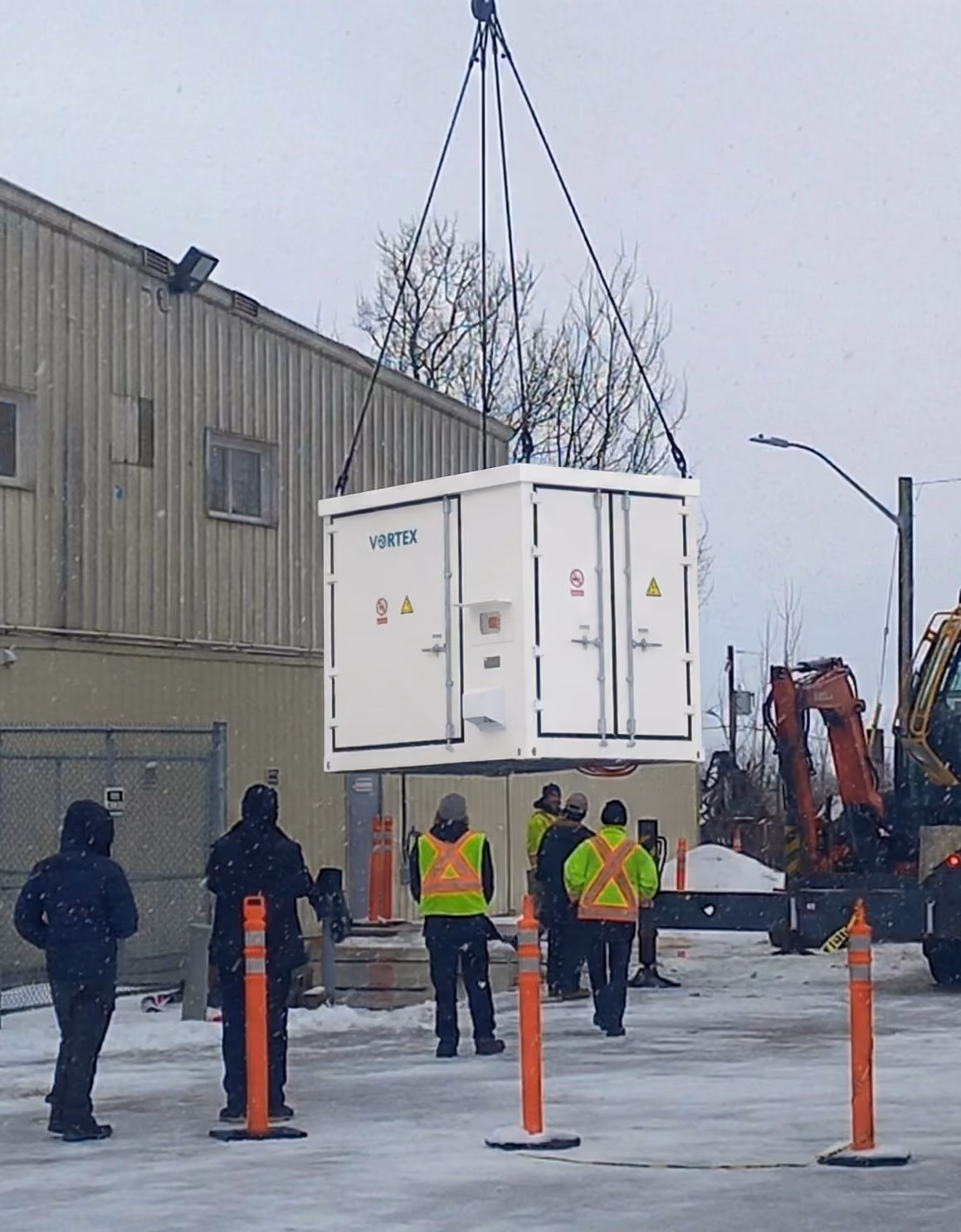A large white container labeled 'VORTEX' is suspended by cables and being lifted at a snowy outdoor industrial site with workers wearing high-visibility vests and winter clothing standing nearby.