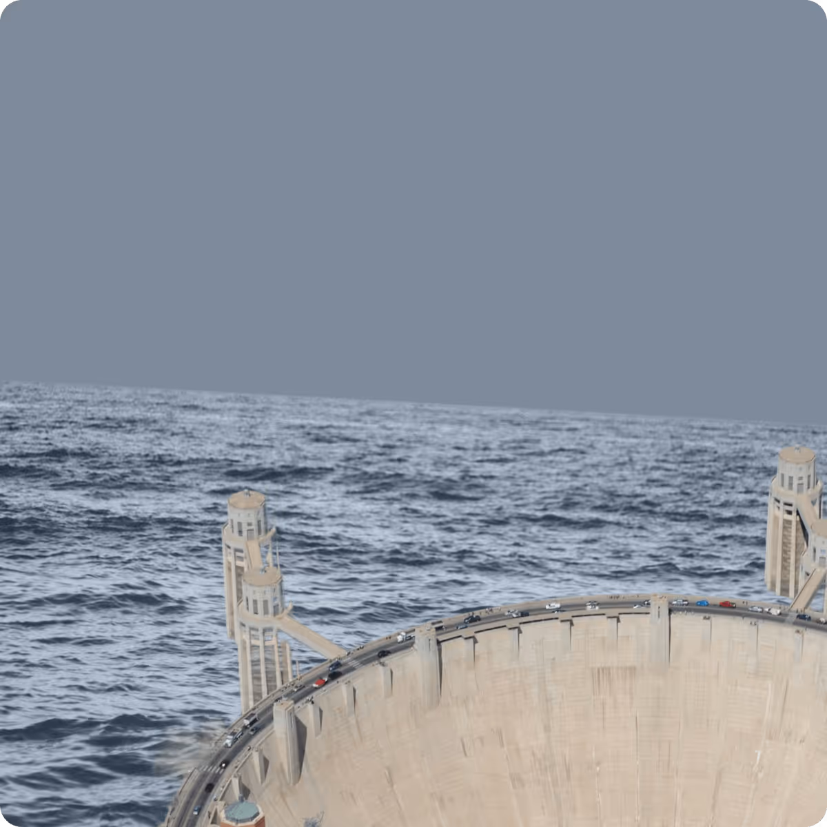 A large curved dam structure with a line of cars driving along the top, set against an open body of water and a cloudy sky.