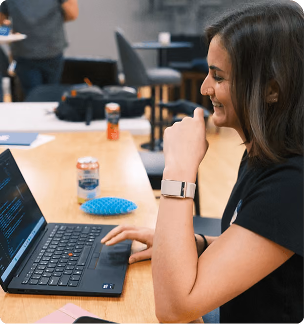 A woman with brown hair and a nose ring sits at a wooden table working on a laptop, smiling while looking at the screen, with a can of drink and a blue textured object nearby.