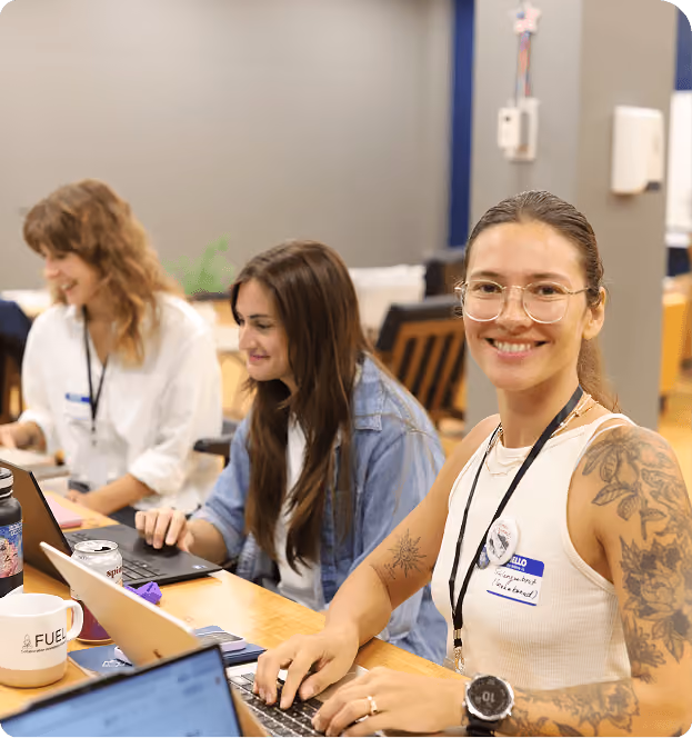 Three women sitting at a shared desk working on laptops, one smiling at the camera wearing glasses and a white sleeveless top with tattoos on her arm.
