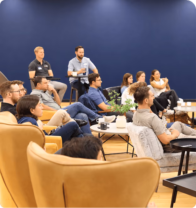 A group of people seated attentively in a modern room with blue walls, sitting on yellow armchairs, gray chairs, and stools, some with drinks, focused on a presentation or speaker outside the frame.