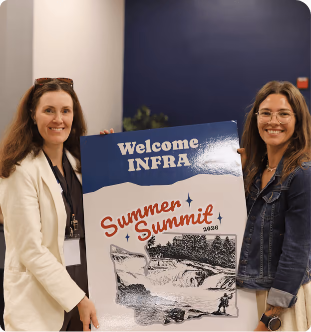 Two women smiling and holding a large poster that reads 'Welcome INFRA Summer Summit 2026' with an illustration of a waterfall and trees.
