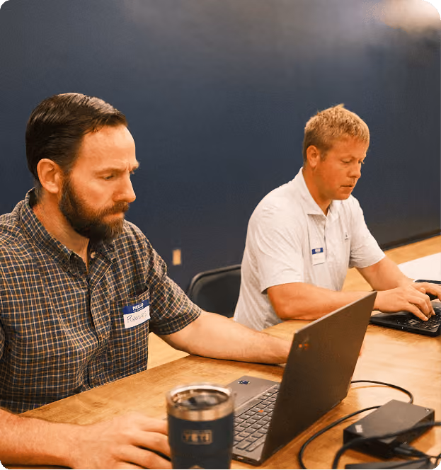 Two men sitting at a table working on laptops; the man on the left wears a plaid shirt with a name tag that reads 'Reese,' and the man on the right wears a white shirt with a name tag. A blue YETI cup is on the table in front of the man on the left.