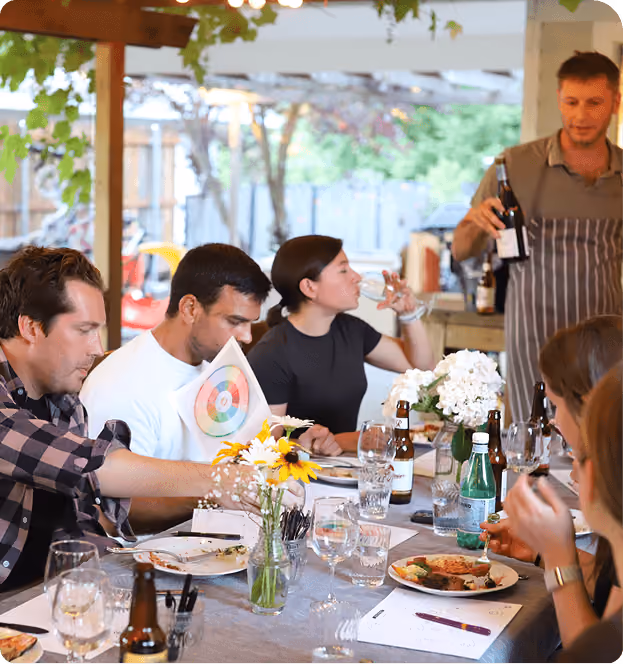 A group of people seated around a dining table outdoors, engaging in conversation and eating; a man in an apron is pouring wine; the table has plates with food, glasses, bottles, and vases of flowers.