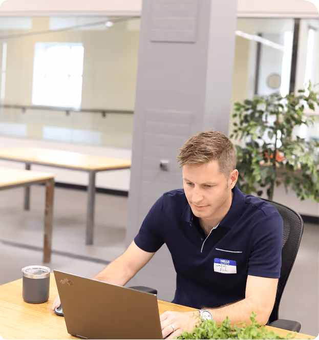 Man wearing a dark blue polo shirt with a name tag sits at a desk working on a laptop with a cup and a plant nearby in a bright, modern office space.