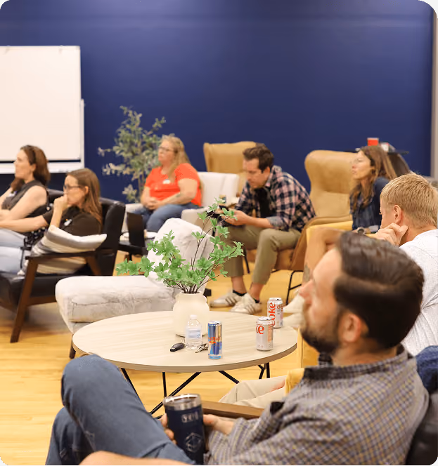 Group of seven people seated in a casual meeting room with a dark blue wall, various chairs, and a round table holding a plant, drinks, and keys.
