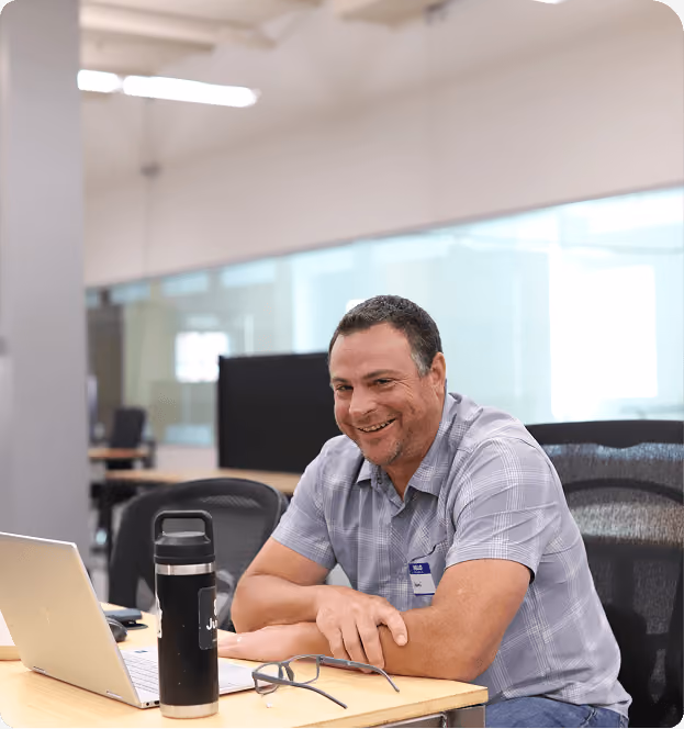 Smiling man with short dark hair wearing a gray checkered shirt seated at a desk with a laptop, black water bottle, and glasses in an office setting.