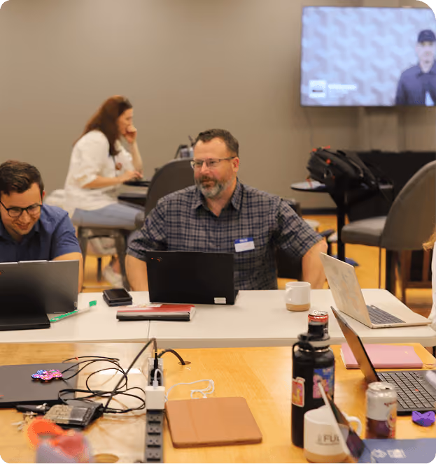 A group of people working on laptops at a table in a casual office setting with a television screen on the wall.