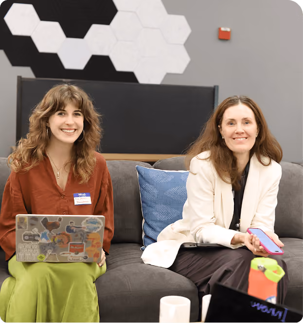 Two women sitting on a gray couch; the woman on the left wears a rust-colored top and green skirt, holding a laptop covered with stickers, while the woman on the right wears a white blazer and dark pants, holding a pink smartphone. They are smiling, with a blue cushion behind them and a wall with hexagonal black and white decorations.