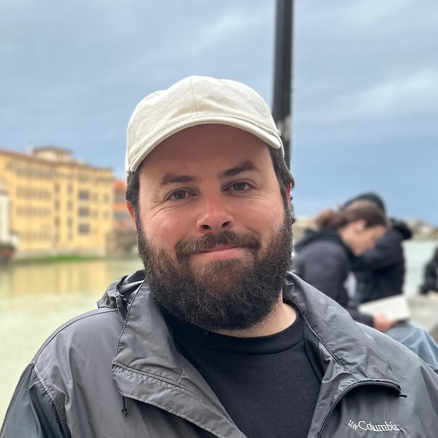 A man wearing a hat and jacket standing in front of a body of water.