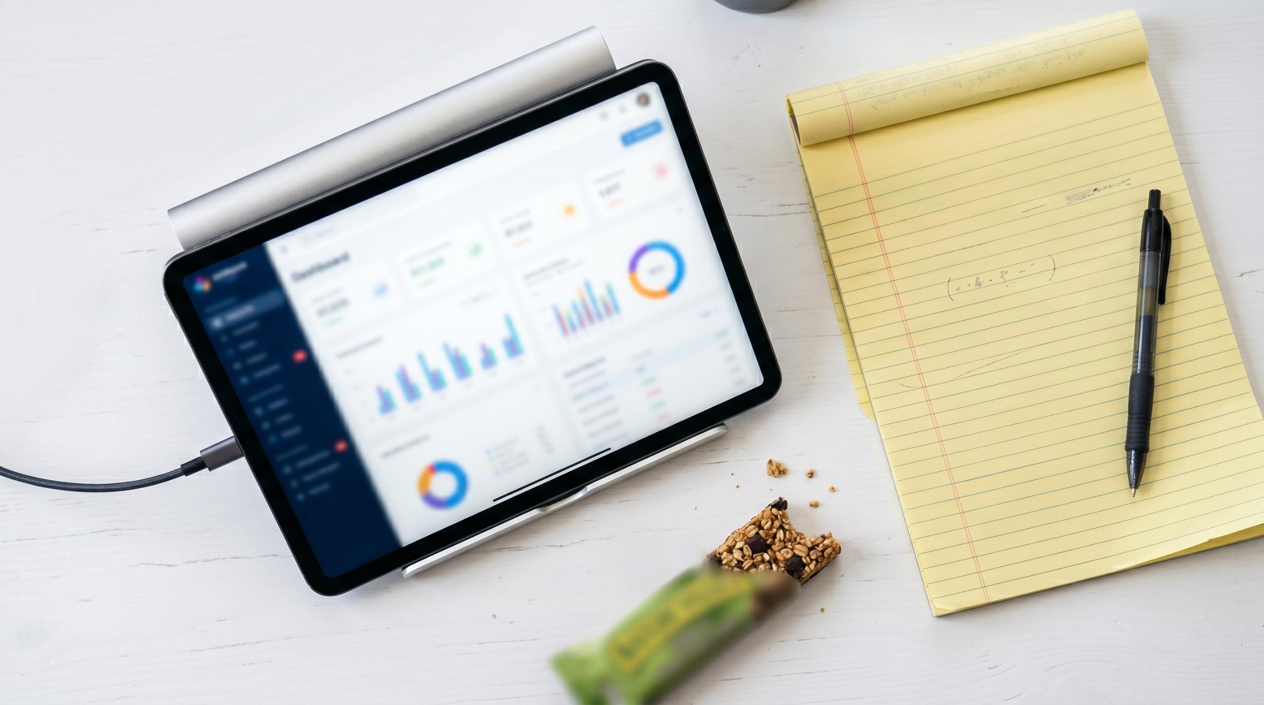 overhead flat-lay photograph of a tablet propped on a stand showing a blurred dashboard interface, surrounded by a clean white desk