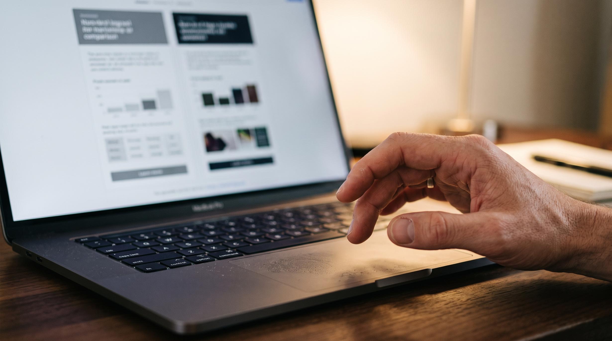 close-up photograph of a hand hovering over a laptop trackpad, screen showing a blurred two-column comparison layout