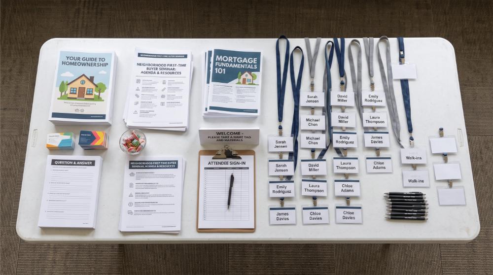 flat-lay photograph of a folding table set up for a neighborhood first-time buyer seminar, printed handouts and name tags arranged on the table