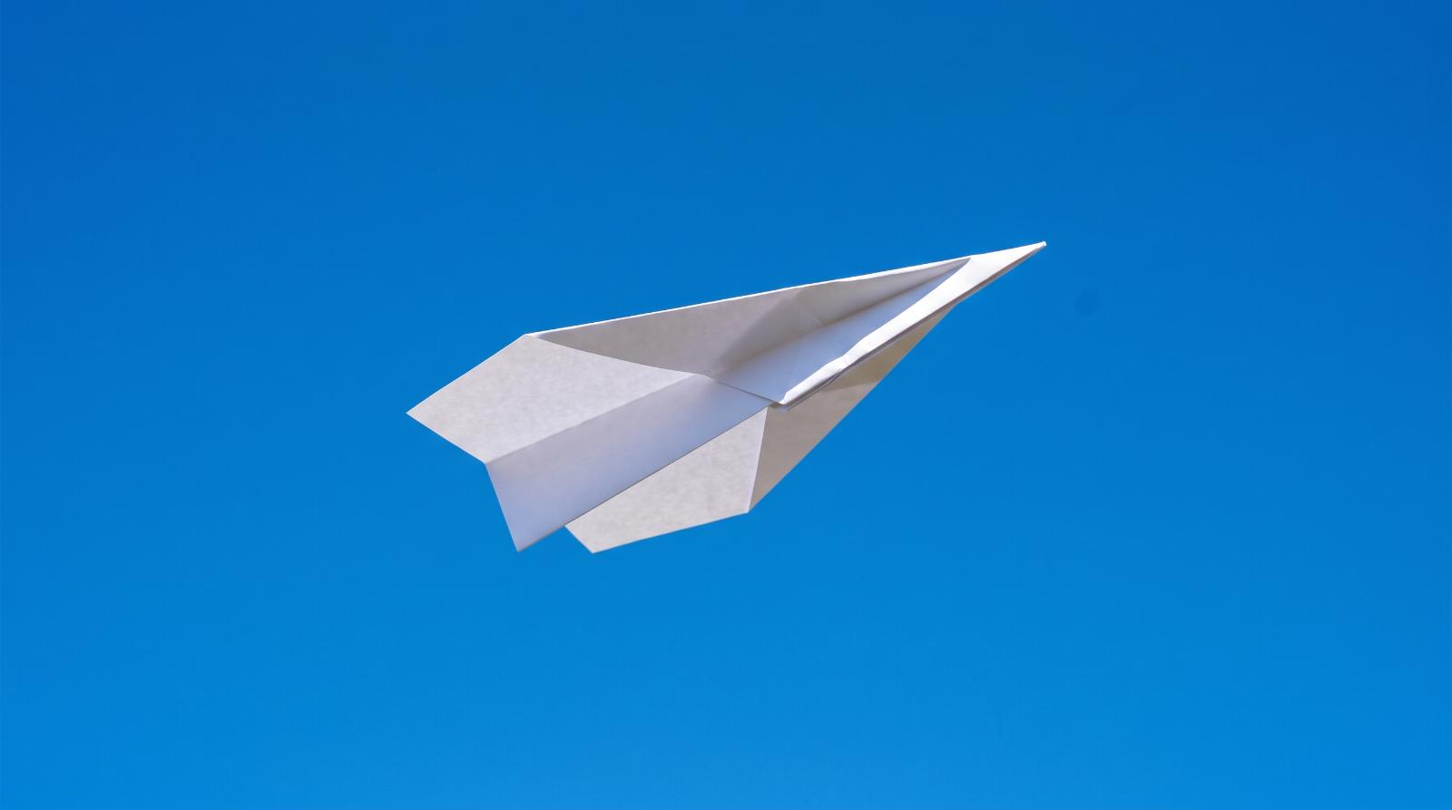a paper airplane mid flight against a bright clear blue sky, sharp deep focus on the paper airplane