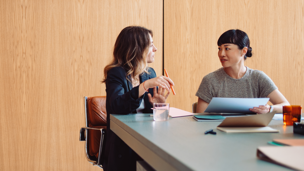 Two women sitting at a table engaged in conversation, with documents and a glass of water on the table.