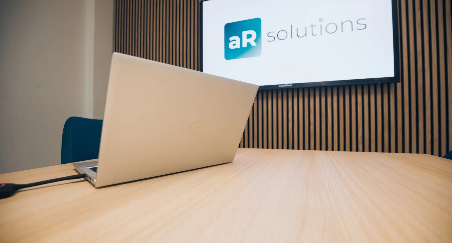 Nahaufnahme eines aufgeklappten HP-Laptops auf einem Konferenztisch, im Hintergrund ein Bildschirm mit dem Logo „aR solutions“ vor einer modernen Holzlamellenwand.
