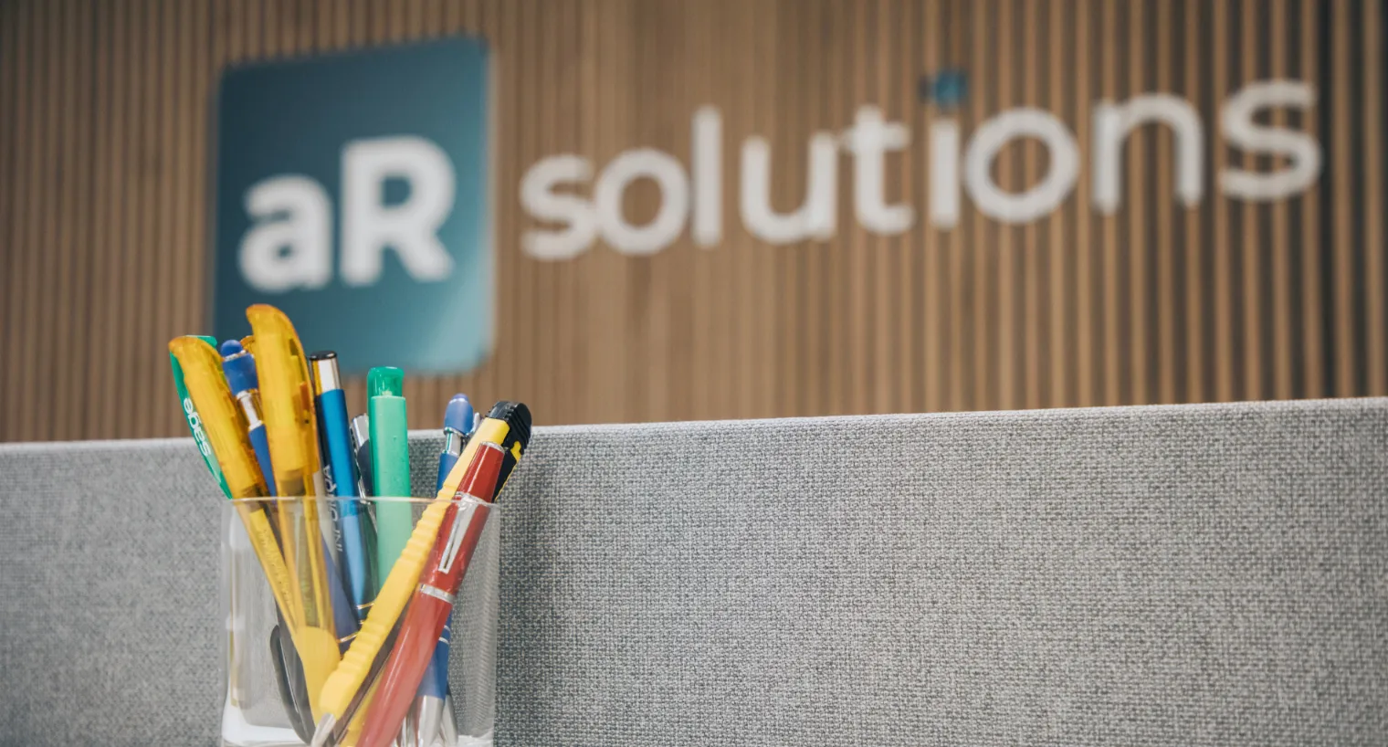 Ein Glas mit bunten Stiften steht auf einer grauen Stellwand im Büro, im unscharfen Hintergrund ist das Logo „aR solutions“ vor einer Holzlamellenwand zu sehen.