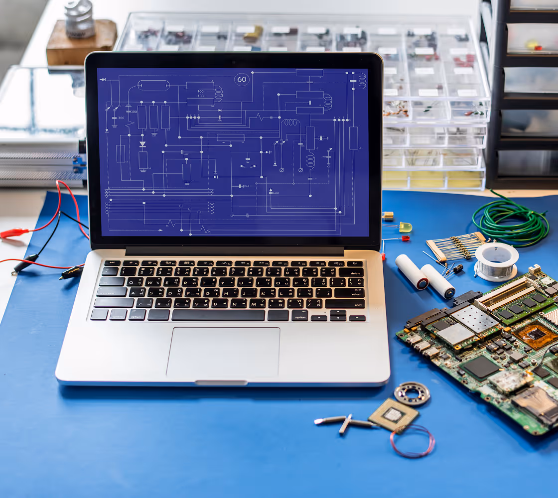 Laptop on a blue workbench displaying an electronic circuit schematic, surrounded by various electronic components and tools.