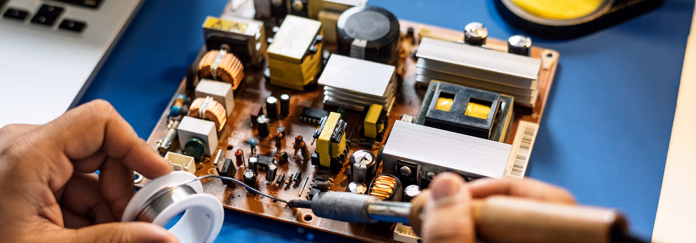 Hands soldering an electronic circuit board with a soldering iron and wire on a blue work surface.