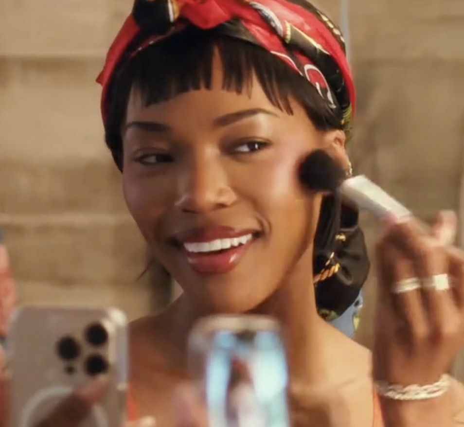 Quen Blackwell smiles while applying makeup in front of a mirror, wearing a red headband and orange top. She holds a phone to record herself, capturing the moment on camera.