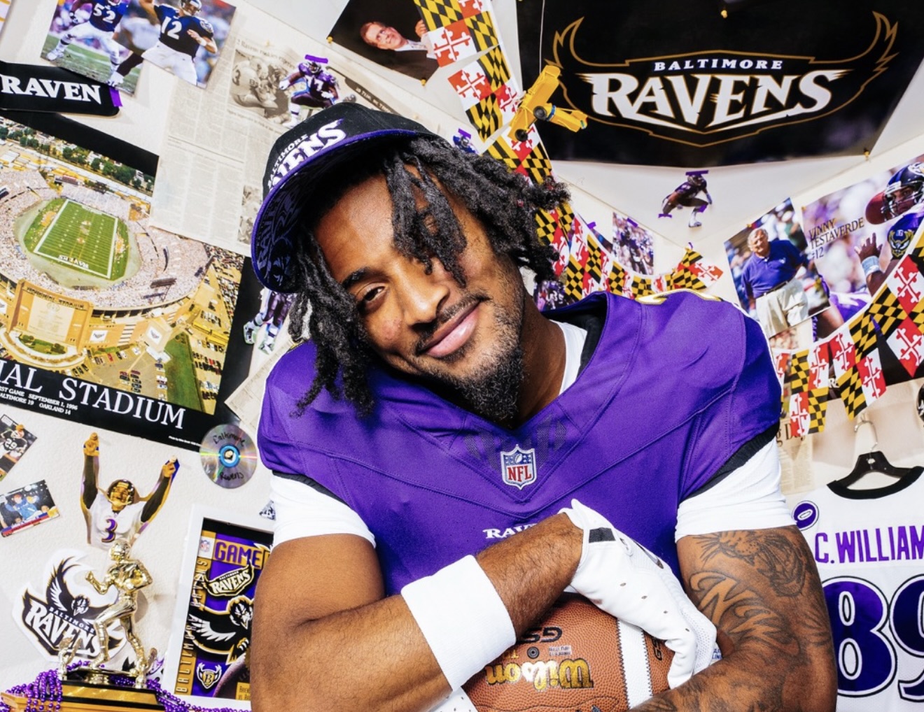 This image shows Lamar Jackson in a Baltimore Ravens uniform, wearing number 8, posing energetically in a room completely covered with Ravens memorabilia. He’s holding a football in one hand and extending his other hand forward as if to stiff-arm an invisible defender. The walls and ceiling are plastered with posters, photos, team logos, and decorations in purple, black, and gold. There are Ravens hats, jerseys, clippings, and even a clock and stereo on a cluttered desk in the corner, making the space feel like the ultimate fan cave. The overall vibe is lively, passionate, and celebratory—capturing both the player’s excitement and the intensity of Ravens fandom.