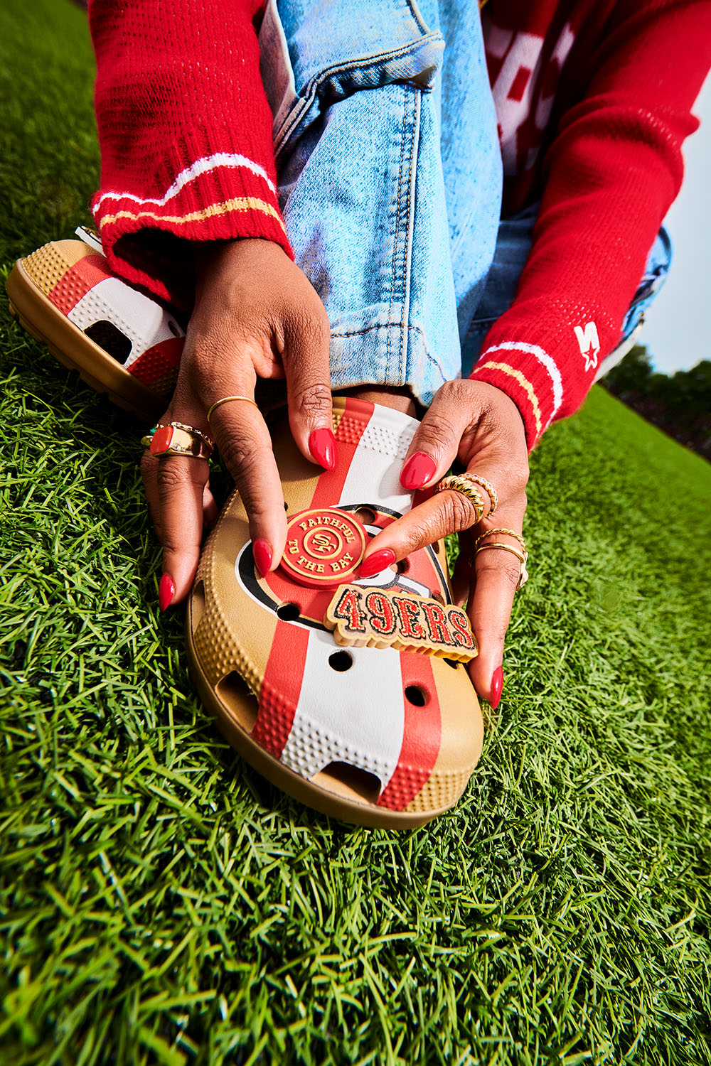 “Close-up of a person wearing San Francisco 49ers–themed Crocs with red, gold, and white stripes, accessorized with gold 49ers charms. The person, dressed in a red sweater and jeans, adjusts the shoe while kneeling on green turf.”