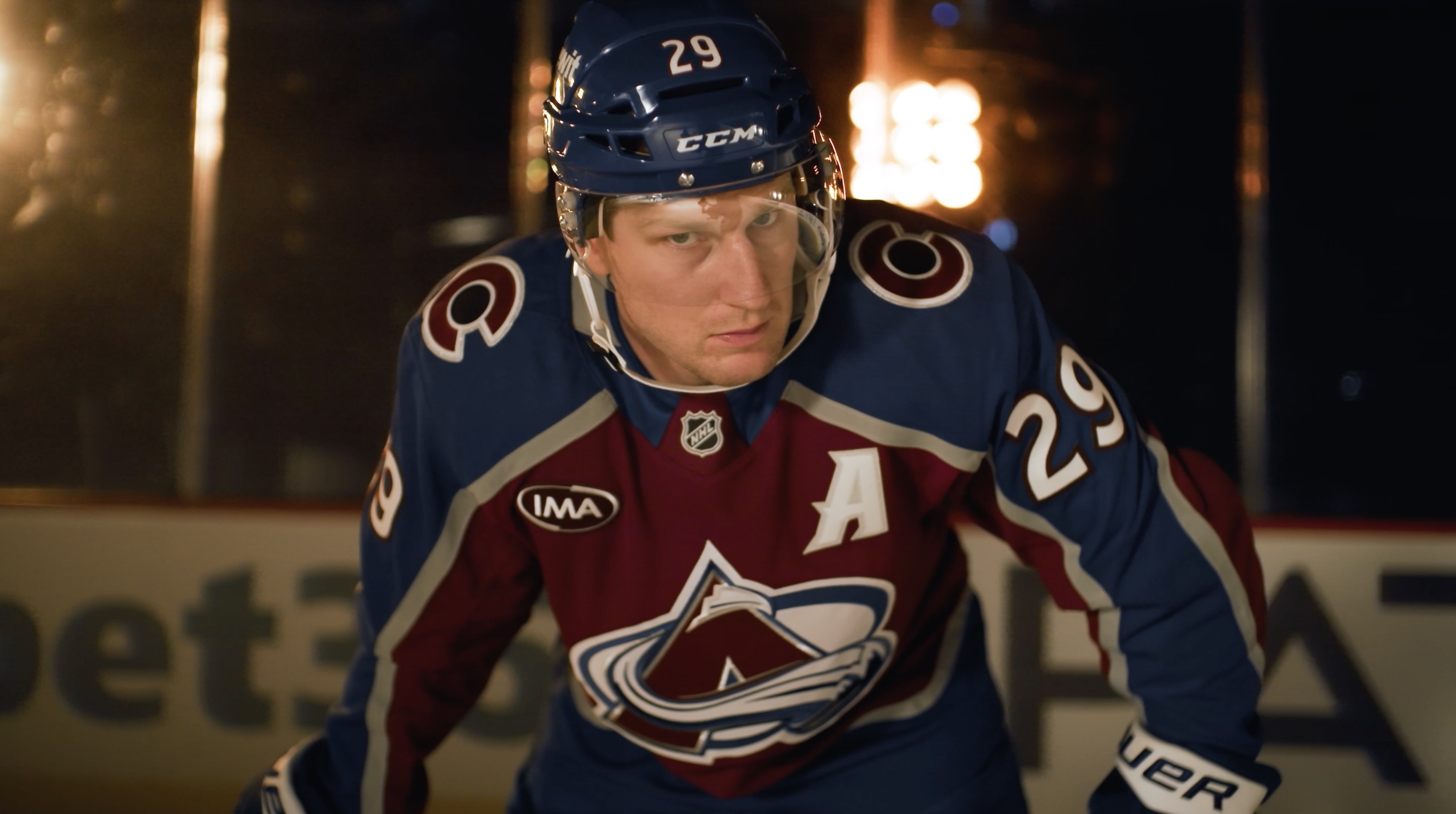 Nathan MacKinnon, suited up in his Colorado Avalanche gear, holds a strong hockey stance while looking up toward the camera with intensity with spotlight grids behind him.
