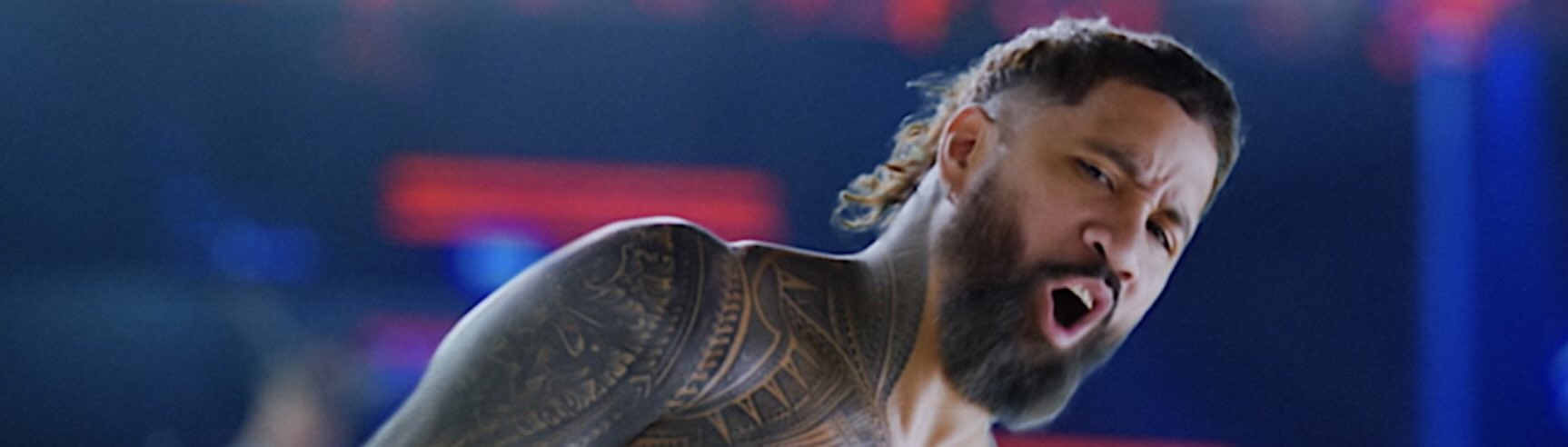 A dramatic shot of a wrestler performing a high energy in ring move, with bright arena lights and a blurred crowd behind him.