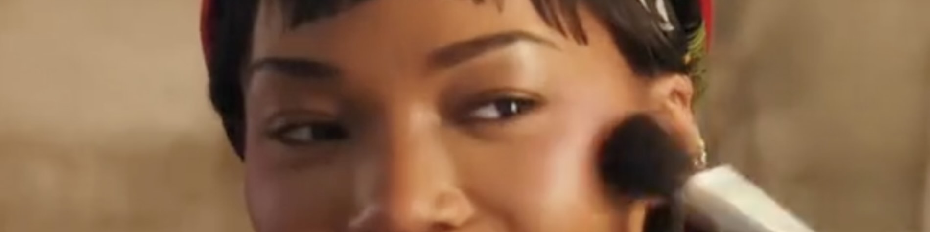 Quen Blackwell smiles while applying makeup in front of a mirror, wearing a red headband and orange top. She holds a phone to record herself, capturing the moment on camera.