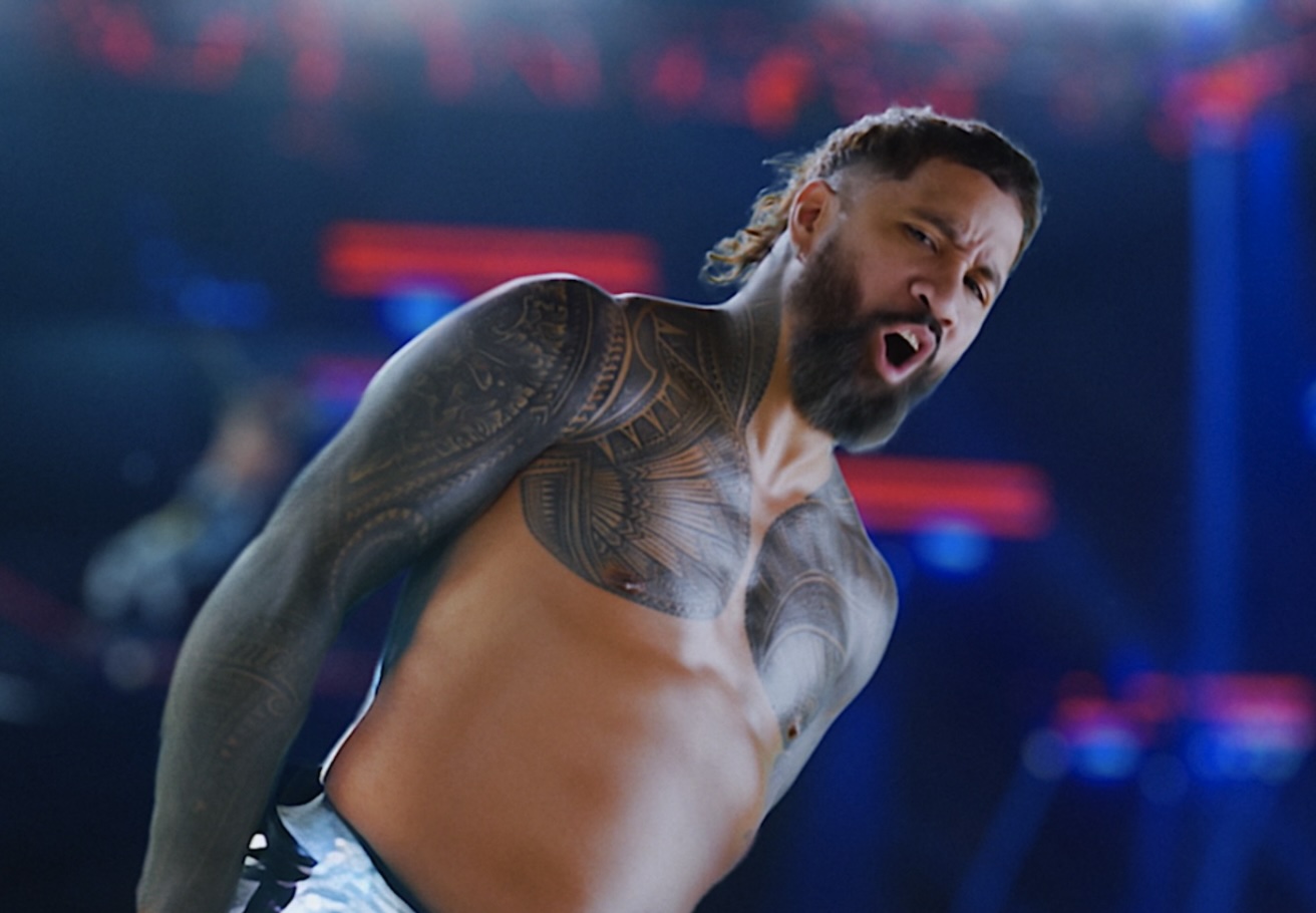 A dramatic shot of a wrestler performing a high energy in ring move, with bright arena lights and a blurred crowd behind him.