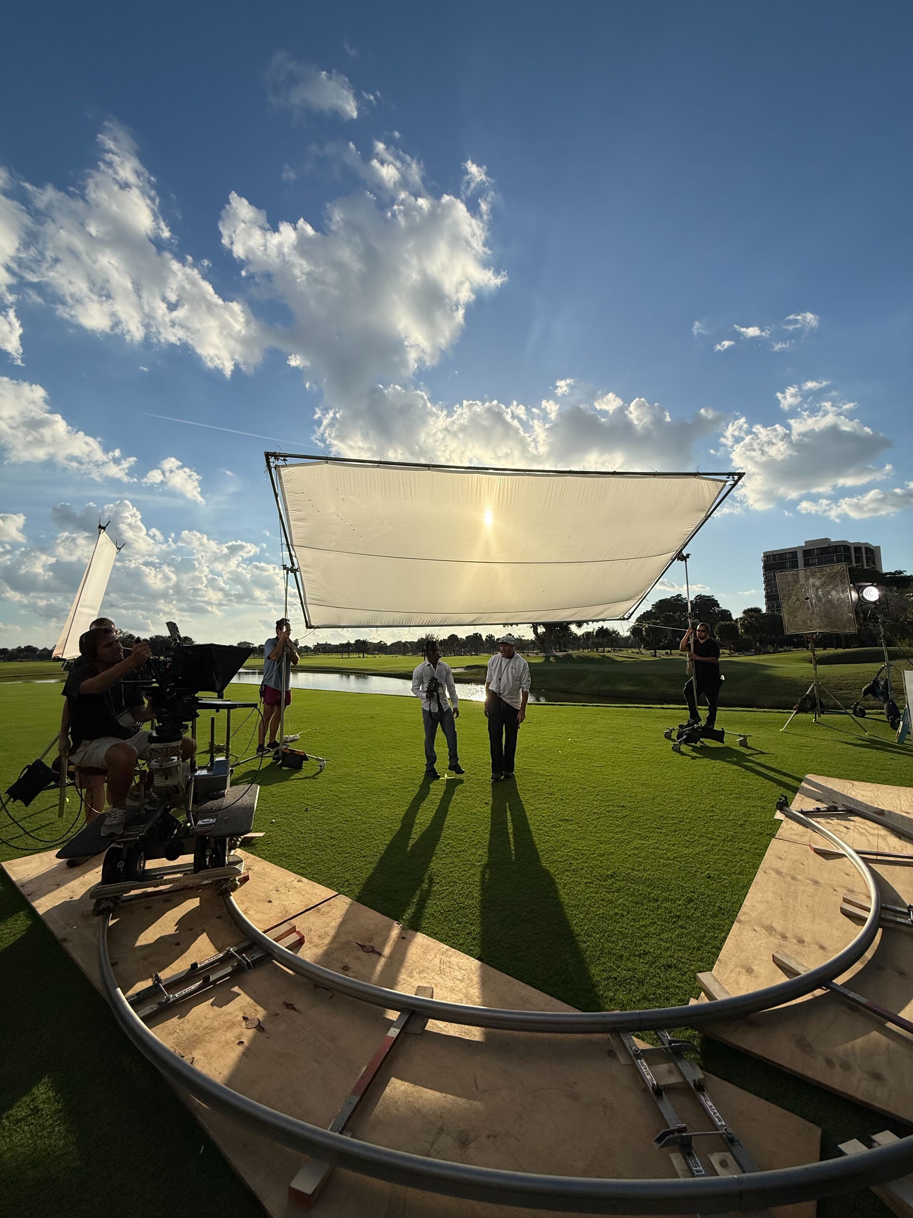 In this BTS shot, three production crew stand around a camera and look at the shot. One person holds a white skrim to reflect light onto the talent.