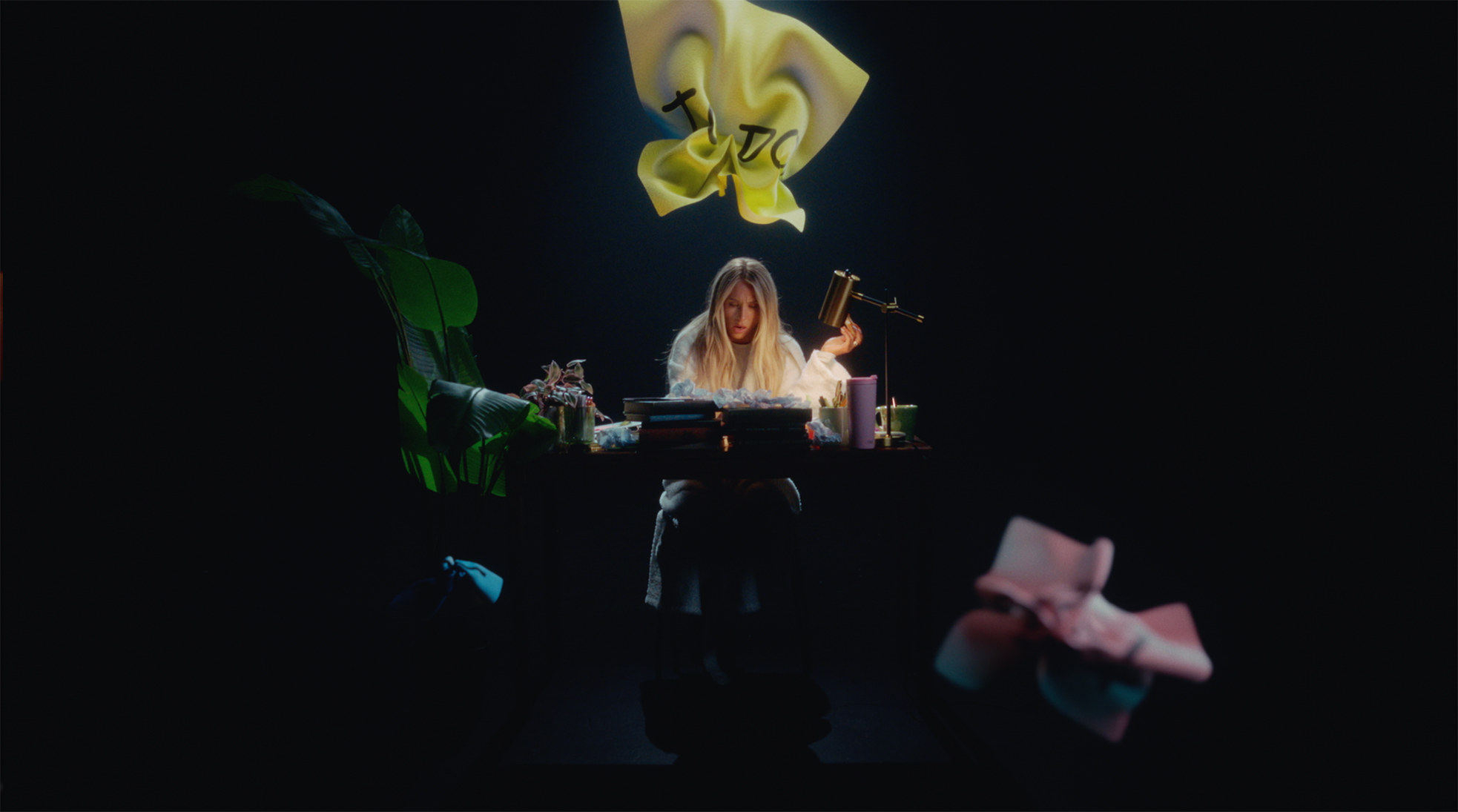Alix Earle in a black dress sits at a wooden desk working on a laptop, surrounded by plants and office supplies. Behind her, a large screen displays a colorful mood board in a PowerPoint presentation.