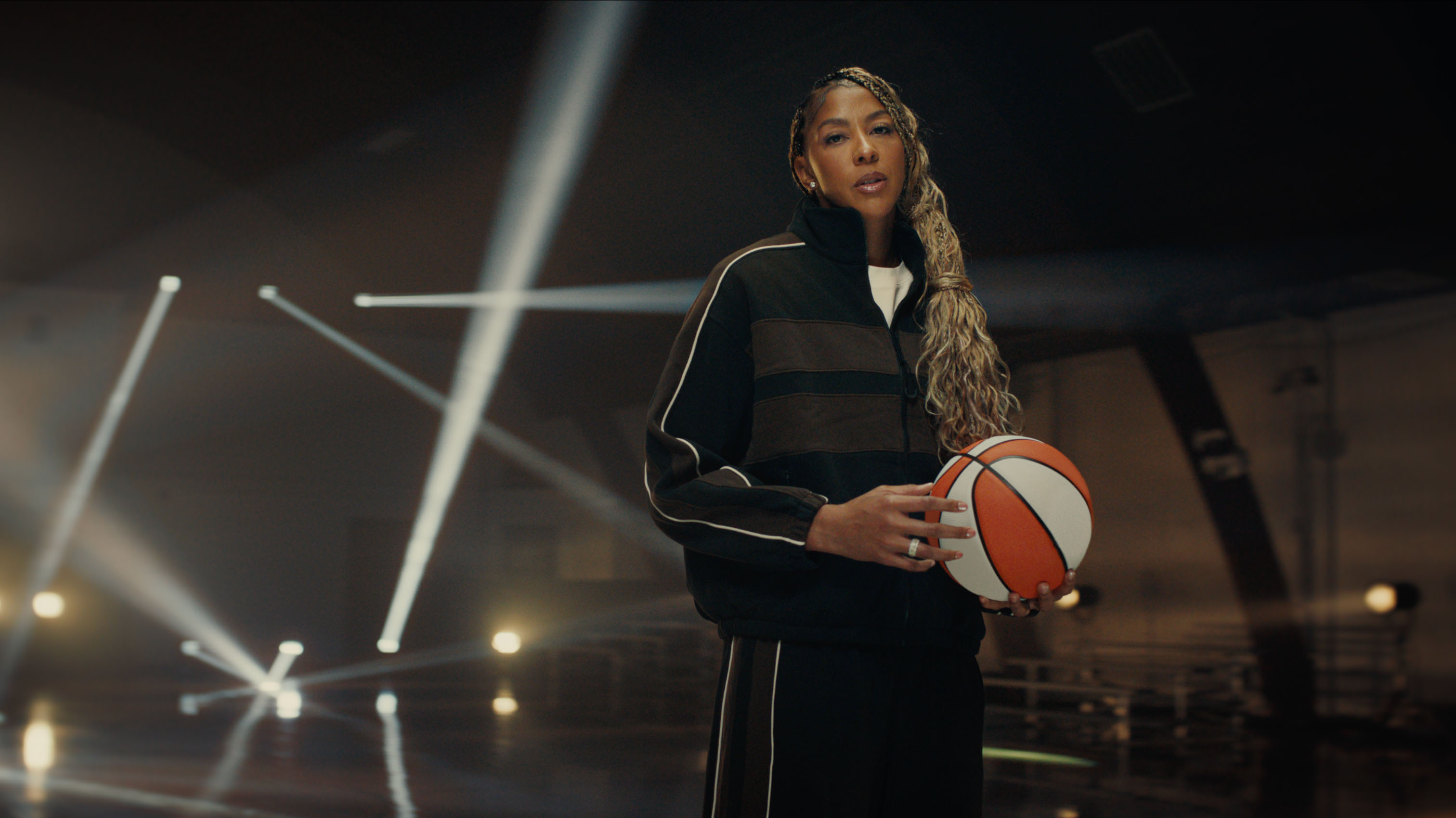 Candace Parker in a black tracksuit stands in a dark indoor basketball gym holding an orange and white basketball. Dramatic beams of light cut through the space behind her as she looks toward the camera.