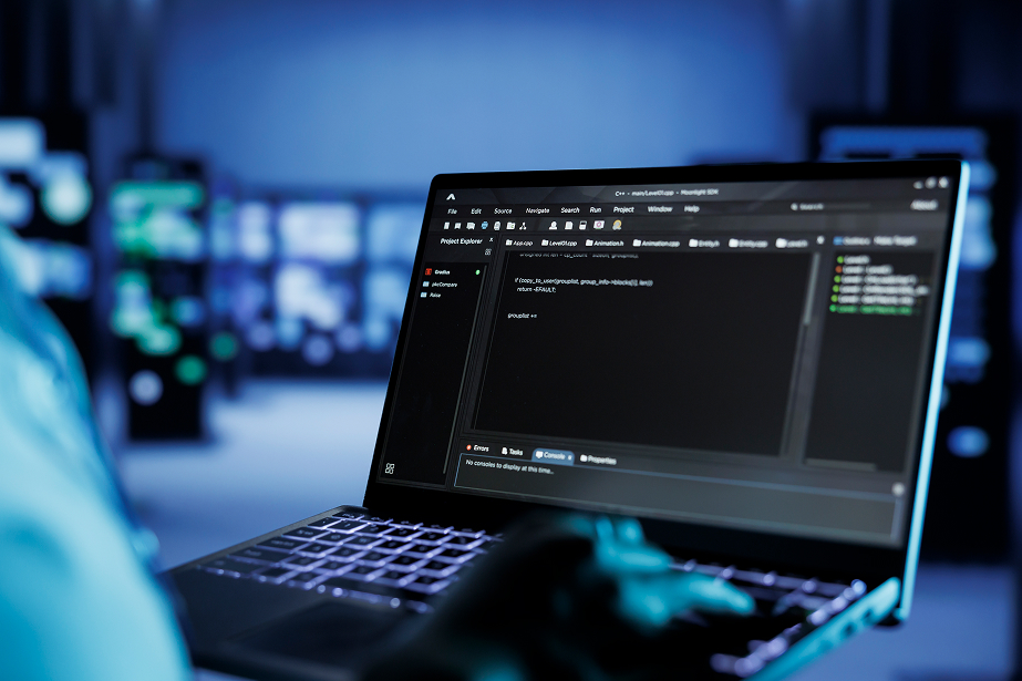 A person using a laptop with code editor software open on the screen in a dark, high-tech environment. The screen displays programming code, and the scene suggests cybersecurity or software development activities.