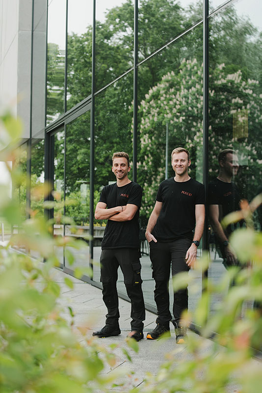 Twee mannen in zwarte werkoutfits staan voor een glazen gebouw met groene bomen op de achtergrond.