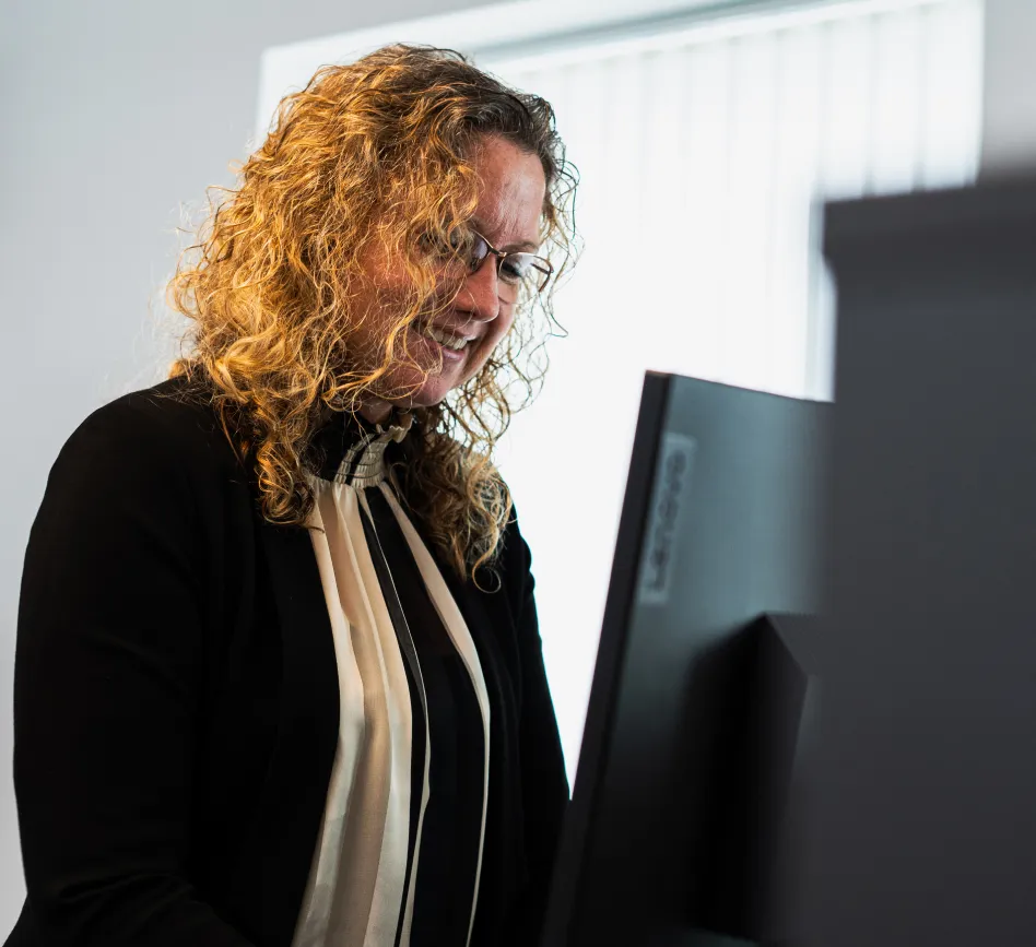 Kvinde med krøllet hår og briller smilende ved en computerskærm i et lyst kontormiljø.
