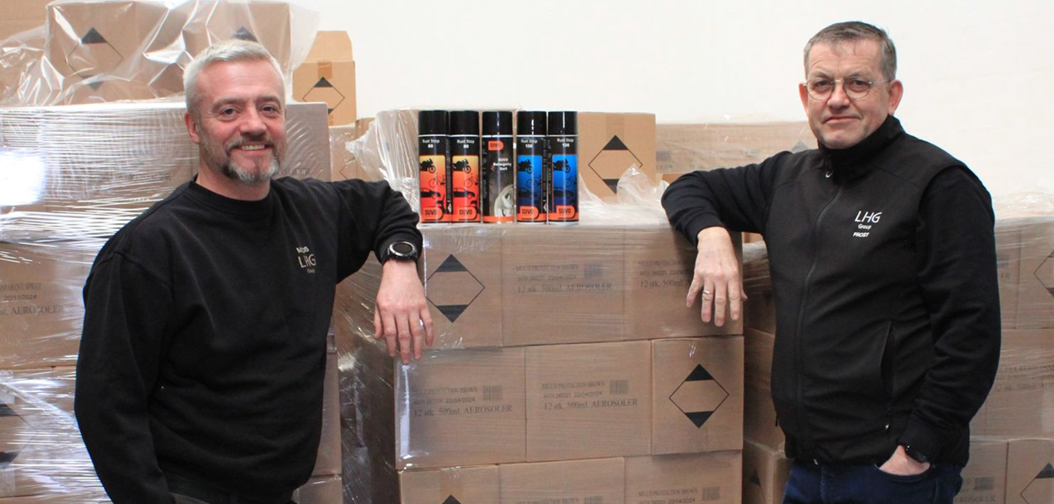 Two men standing with an arm on a pallet of bokses with some aerosol cans on top