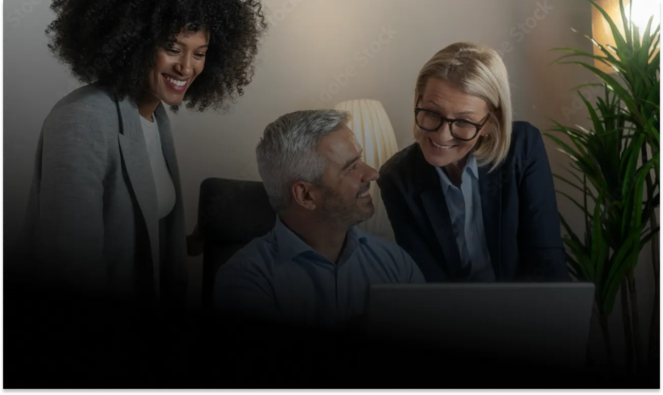 Three coworkers smiling and discussing work around a laptop in an office setting with a lamp and plant in the background.