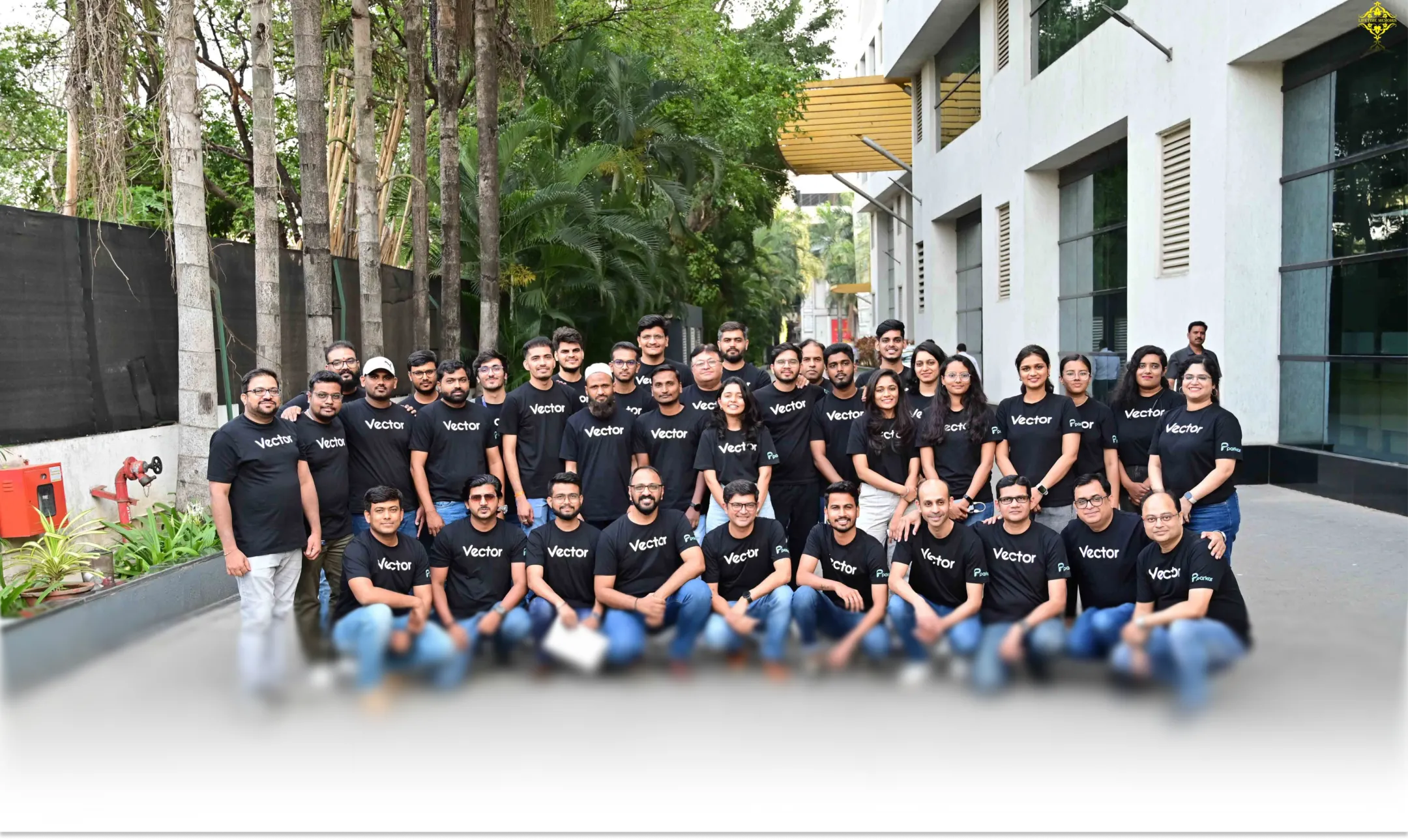 Large group of men and women wearing matching black 'Vector' t-shirts posing outdoors in front of a building and greenery.