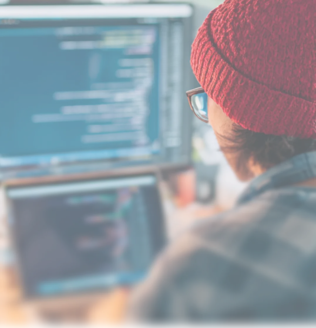 Person wearing a red knit hat and glasses working on a laptop with code and graphs displayed on a screen in the background.