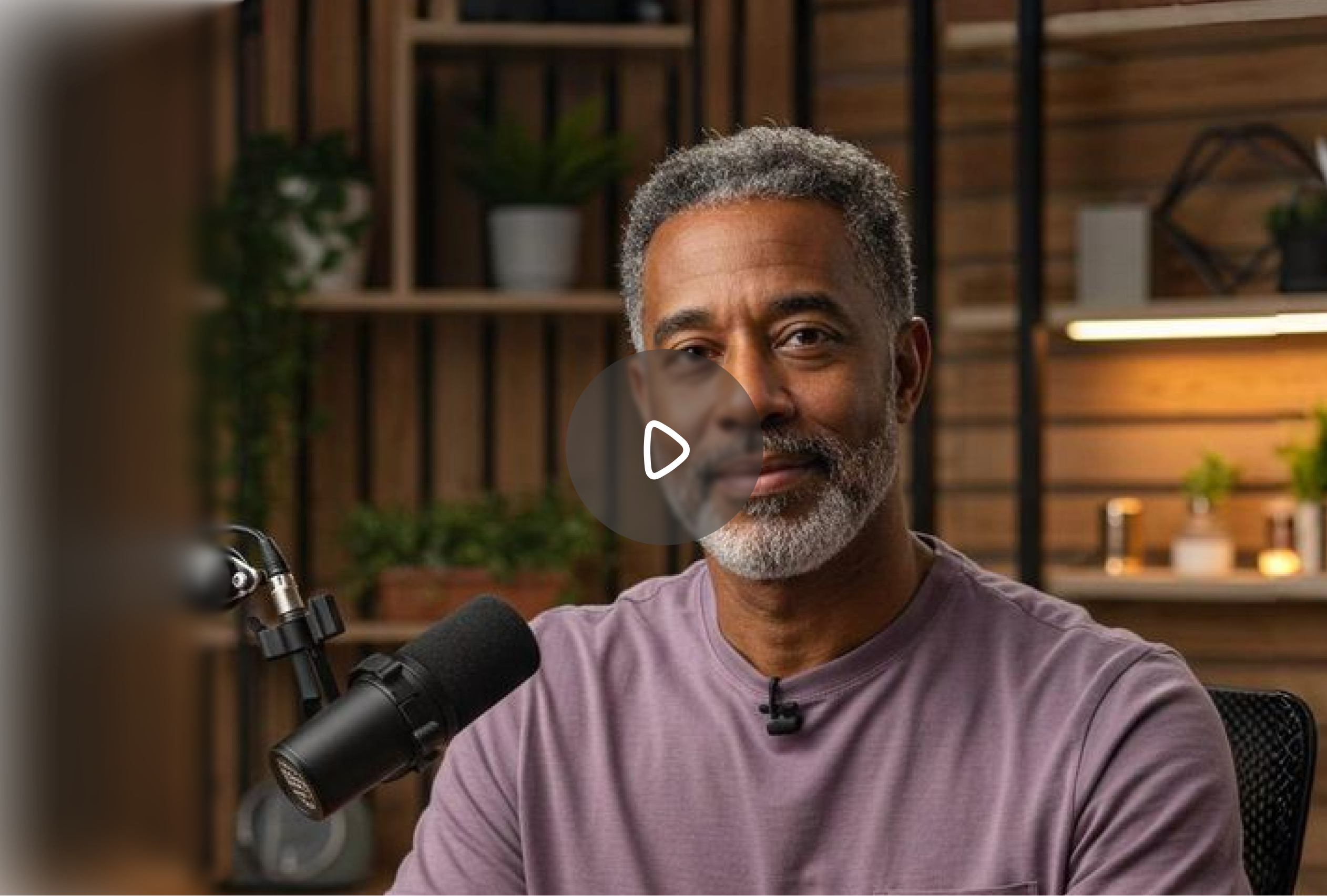 Middle-aged man with gray beard speaking into a microphone in a cozy, well-lit room with plants and wooden shelves.